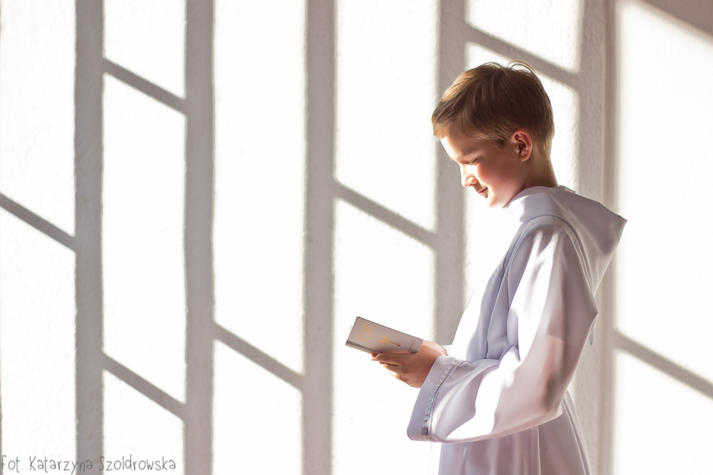 fotografia komunijna przedstawiająca chłopca czytającego książeczkę do nabożeństwa we wnętzru spowitymjasnym światłem. Zdjęcie wykonano w Krakowie Mistrzejowicach w kościele św. Maksymiliana Marii Kolbego