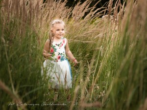 Fotografia kilkuletniego dziecka. Jasnowłosa Dziewczynka w białej sukience w kwiaty stoi pośród kwitnących traw. Sesja dziecięca wykonana w plenerze w Krakowie Mistrzejowice, Nowa Huta