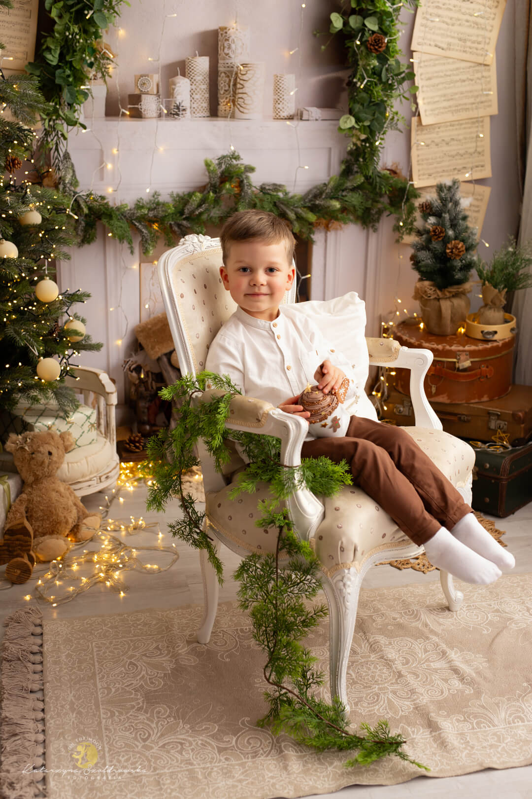 minisesja świąteczna Kraków. Sesja świąteczna w studio w Krakowie. Mały chłopiec siedzi na eleganckim jasnym fotelu na tle kominka i choinki