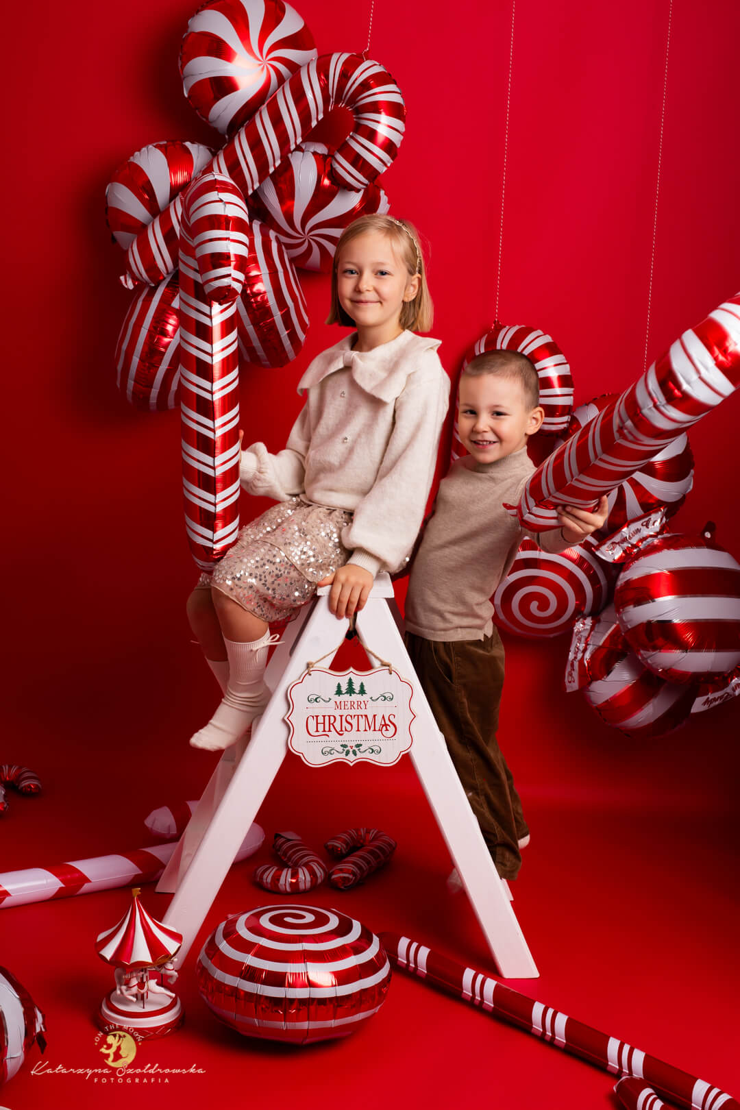 Sesja świąteczna Krakó. Secja rodzeństwa w studio w Krakowie na czerwonej nowoczesnej scence z balonami w kształcie świątecznych cukierków