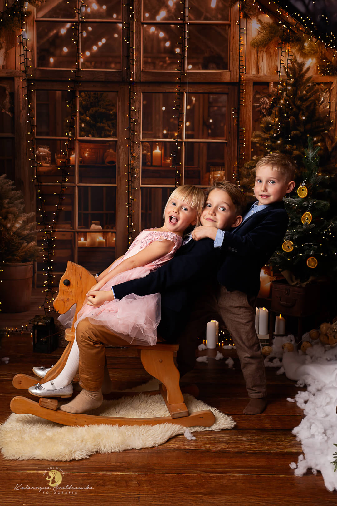 Sesja świąteczna Kraków. Zdjęice trójki rodzeństwa. Roześmiane dzieci siedzą na bujanum konuku na świątecznej scence w kolorze brązowym ze światełkami