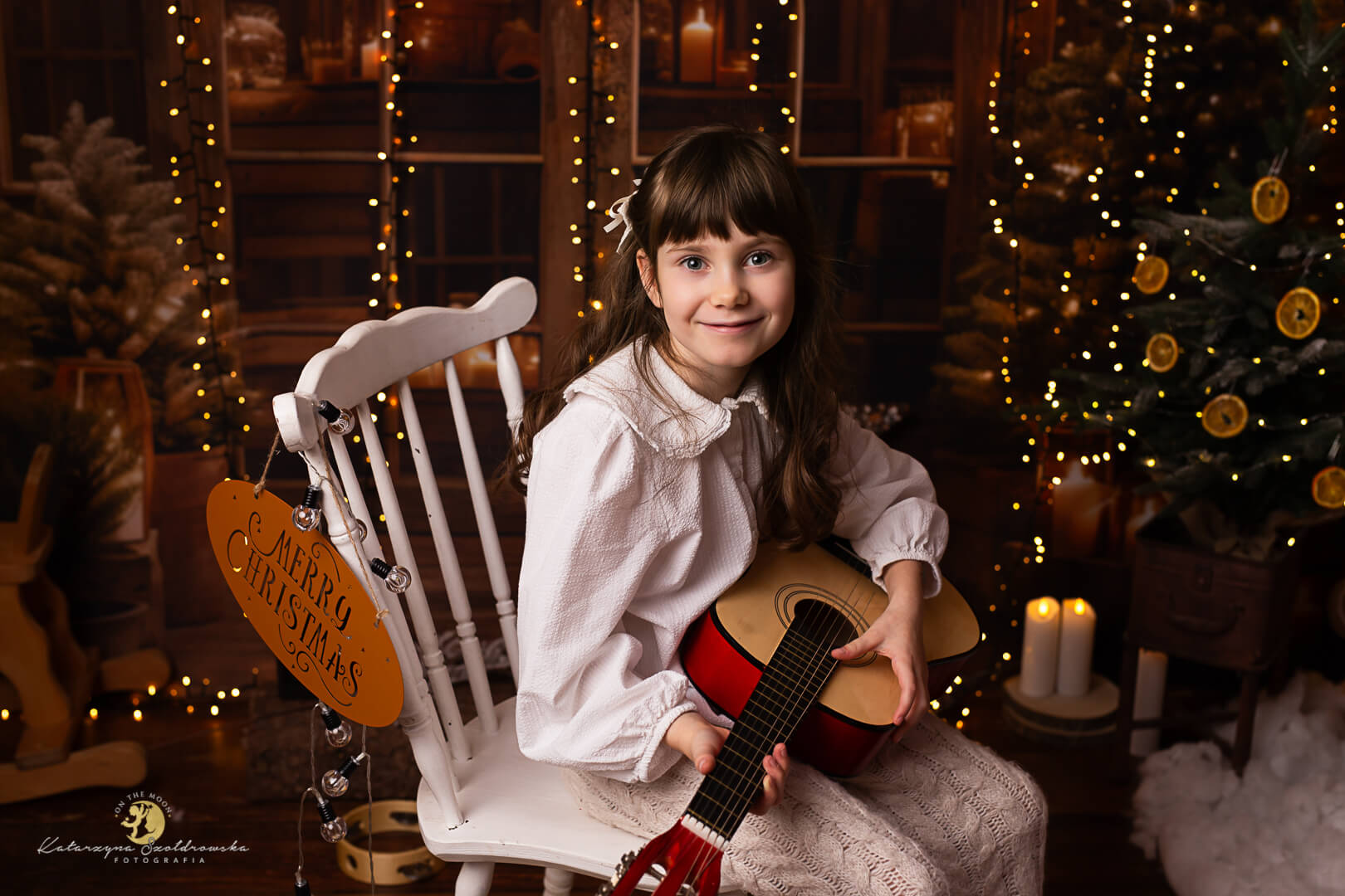 sesje świąteczne Kraków. Swiąteczne zdjęcie dziewczynki grającej na gitarze na brązowej scence ze światełkami. Zdjecie z sesji świątecznej w studio w Krakowie w roku 2025