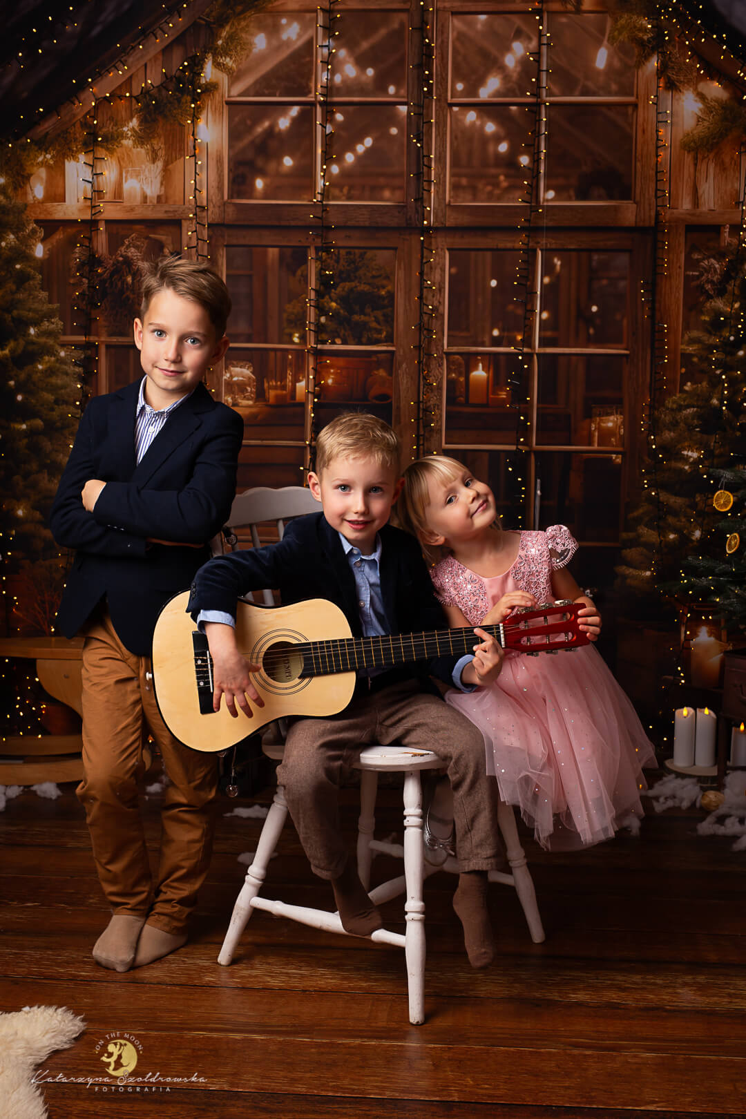 sesje świąteczne kraków2025. Zdjęice rodzensta na brązowej świątecznej scence ze światełkami. Chłopiec gra na gitarze